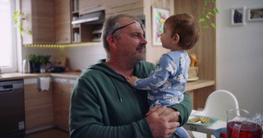 Baba kucağında bebek ve gülümsüyor. Sıcak bir mutfak ortamında neşeli ve sevecen bir an yaşıyor.