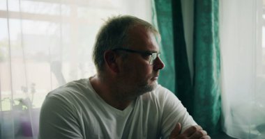 Pencere kenarında oturan bir adam, düşünceli bir ifadeyle dışarı bakıyor, kollarını kavuşturmuş, rahat bir duruş içinde, sakin bir iç mekanda yumuşak perdelerle çevrili.