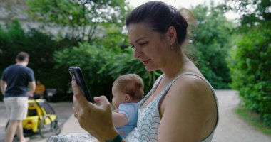 Anne akıllı telefonunu açık havada kullanırken bebeği kucağına alıyor, sonra da kaldırıyor, çocuğuyla yeşil bir park ortamında etkileşime geçiyor.