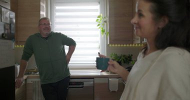 İki yetişkin modern bir mutfakta neşeli bir şekilde sohbet ediyorlar. Mavi bir kupa tutan ve gülümseyen bir kadın. Tezgaha yaslanan bir adam, rahat bir atmosfer.