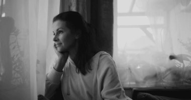 Pencereden dışarı bakan kadın, düşünceli bir duruşla çenesini dinlendiriyor, bitkilerle ve perdelerle çevrili huzurlu bir evde oturuyor, monokromatik tonları yansıtıyor.