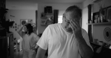 Tartışma sırasında yoğun ifadeli erkek portresi, oturma odasında arka planda kadın, duygusal gerilim ve gergin ilişki dinamiklerine odaklan, siyah-beyaz