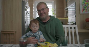 Babam yemek masasında kucağında bebeğini tutarken gülümsüyor, sıcak ve sevgi dolu bir aile anı yaratıyor.
