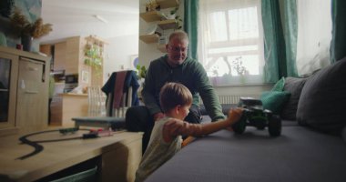 Babası gülümserken oğlu kanepede yeşil bir oyuncak kamyonla oynuyor. Sıcak ve rahat bir oturma odasında, ahşap mobilyaları ve canlı bir iç mekanı var.