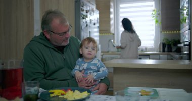 Babam yemek masasında renkli yemek ve içeceklerle bebeği tutarken annem de mutfakta, sıcak aile ortamında sıcak aile ortamını hazırlıyor.