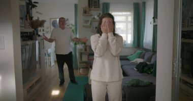 Kadın, arka planda bir erkekle tartışırken kararlı bir şekilde oturma odasında duruyor. Öfkeyle el kol hareketi yapıyor, bir çift ilişkisindeki gerginliği ve çatışmayı yakalıyor..