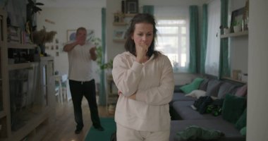 Kadın, arka planda bir erkekle tartışırken kararlı bir şekilde oturma odasında duruyor. Öfkeyle el kol hareketi yapıyor, bir çift ilişkisindeki gerginliği ve çatışmayı yakalıyor..