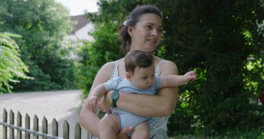 Anne dışarıda bebeğini tutuyor, çocuğuyla meşgul olurken gülümsüyor, arka planda yeşillikler ve ağaçlar var, doğal park ortamında neşeli ve hassas bir an.