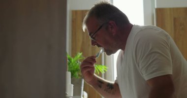 Banyo lavabosunda dişlerini fırçalayan adam, yoğun ifadeli yan profil görüntüsü, parlak ve temiz ev ortamında sabah diş bakımı rutini