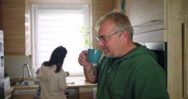 Annesi buzdolabını kollarında bebekle kontrol ederken elinde bardak tutan bir adam, tipik bir sabah rutini sırasında sıradan bir aile anı, sıcak ve besleyici ebeveynlik sahnesi.