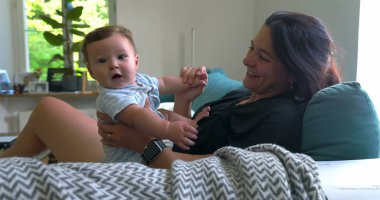 Anne ve bebek kanepede kaynaşıyor, bebek gülümsüyor ve meraklı görünüyor, sakin ve sıcak bir aile anının tadını çıkarıyor.