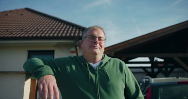 Evin önündeki konut kapısına yaslanan adam, gülümseyen ve rahat, banliyö yaşam tarzı ve mülk çevresi odaklı