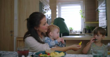 Anne kahvaltıda çocuklarla etkileşime giriyor, bebek yemek yerken, kardeş yemek yiyor, sıcak bir aile ortamında anne-baba iletişimi kuruyor.