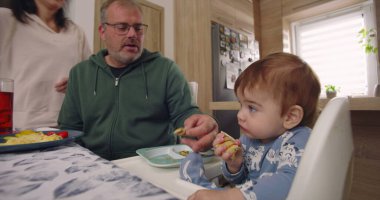 Baba yemek sırasında bebek maşasında oturur, anne tabağın yanında durur, aile bağlarını yemek masasında kurar, çocuk yemek yer.