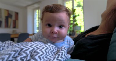 Bebek battaniyeye uzanmış merakla bakarken anne arka planda doğal ışık ve sıcak aile atmosferiyle dolu rahat bir oturma odasında gülümsüyor.