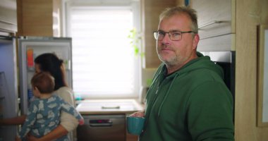 Annesi buzdolabını kollarında bebekle kontrol ederken elinde bardak tutan bir adam, tipik bir sabah rutini sırasında sıradan bir aile anı, sıcak ve besleyici ebeveynlik sahnesi.