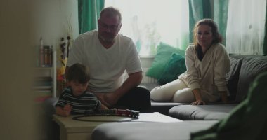 Anne ve baba kanepede oturup çocuğa gülümsüyor kahve masasında oyuncak trenle oynuyor rahat oturma odası düzeninde, neşeli aile anları, kapalı alanda kaynaşma zamanı.