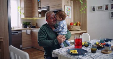 Baba kucağında bebek ve gülümsüyor. Sıcak bir mutfak ortamında neşeli ve sevecen bir an yaşıyor.