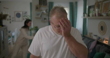 Oturma odasında hararetli tartışmalar yaşayan bir çift, görünürde üzgün duran bir adam, arka planda duygusal hareketler yapan bir kadın, ilişki çatışması sırasında gergin bir an.