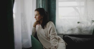 Kanepede öne doğru eğilen kadın, elinde çenesiyle pencereden dışarı bakıyor, sessiz bir ev ortamında bitkilerle ve perdelerle çevrili, sakin bir tavırla yansıyor.