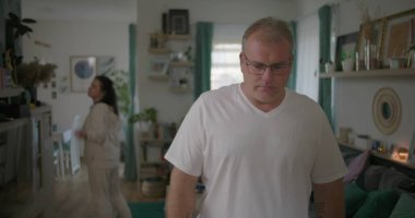 Erkek, arka plandaki kadınla tartışırken gözle görülür şekilde üzgün, oturma odasında ilişki gerilimi, duygusal ve dramatik anlaşmazlık anı