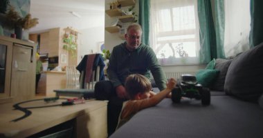 Babası gülümserken oğlu kanepede yeşil bir oyuncak kamyonla oynuyor. Sıcak ve rahat bir oturma odasında, ahşap mobilyaları ve canlı bir iç mekanı var.