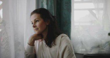 Kanepede öne doğru eğilen kadın, elinde çenesiyle pencereden dışarı bakıyor, sessiz bir ev ortamında bitkilerle ve perdelerle çevrili, sakin bir tavırla yansıyor.