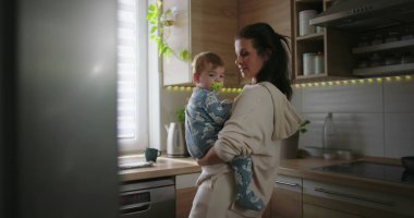 Anne mutfak tezgahının yanında bebeği tutuyor, çocuğa gülümsüyor ve bağ kuruyor, günlük aile rutini sırasında sıcak ve şefkatli ebeveyn anlarını yaşıyor.