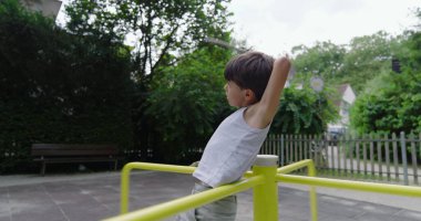 Çocuk sarı bir atlıkarıncada dikilirken elinde sopayla arka planda yeşillik ve çitle