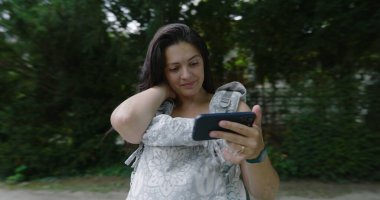 Anne, bebeğini parktaki bir taşıyıcıda taşırken akıllı telefon kullanıyor. Serin yeşil bir ortamda günlük bir yürüyüş sırasında açık havada birden fazla iş yapıyor.