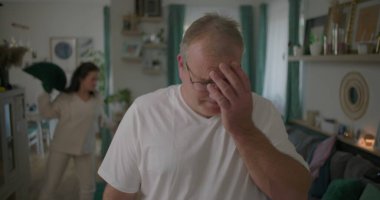 Oturma odasında hararetli tartışmalar yaşayan bir çift, görünürde üzgün duran bir adam, arka planda duygusal hareketler yapan bir kadın, ilişki çatışması sırasında gergin bir an.