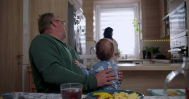 Baba bebeğini kucağında tutarken anne mutfakta hazırlanıyor sıcak ve sevgi dolu bir aile ortamında ev ortamında.