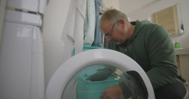 Çamaşır makinesinin önünde diz çökmüş bir adam, temiz havluları ve parlak döşeli duvarları olan modern bir banyoda çamaşırları dikkatlice yüklüyor.