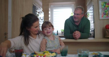 Anne, büyük çocukla yemek masasında gülerken, baba mutfak tezgahından sıcak bir şekilde izliyor, sıcak bir ev ortamında neşeli bir aile anı.