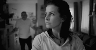 Kadın hararetli tartışmalar sırasında erkeklerden yüz çeviriyor, mesafeli ve üzgün görünüyor, erkek ise arka planda hayal kırıklığıyla işaret ediyor, duygusal gerilimi ve çatışmayı yakalıyor.