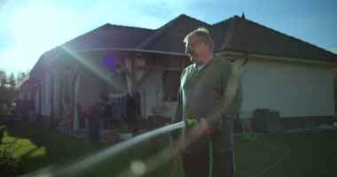 Arka planda bahçe hortumu, konut evi ve avlusu olan bahçeyi sulayan bir adam, banliyö evlerinde yeşil ve güneş ışığı olan rahat ve rahat bir an.