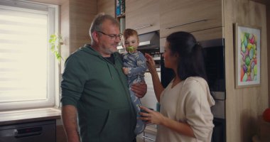 Anne, modern mutfakta babayla konuşurken bebeğin kafasına şefkatle dokunuyor. Günlük ev ortamında sıcak aile etkileşimi.