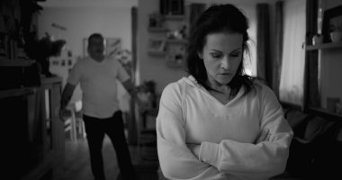 Tartışma sırasında kollarını çapraz uzatmış bir kadın, arka planda yürüyen ve el kol hareketi yapan bir adam, gergin ve duygusal ilişki çatışması, siyah ve beyaz