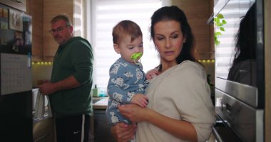 Anne kucağında bebek, baba arka planda çalışırken aile dinamiklerini ve günlük rutinlerini gözler önüne seriyor.