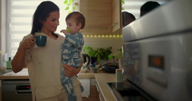 Anne mutfakta bebeği tutarken çocuk sıcak fincana uzanır, günlük aile rutini sırasında koruyucu ebeveynlik anı, sıcak bir evde bakım ve güvenlik