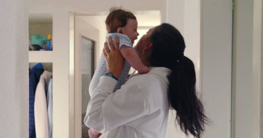 Anne koridorda bebeğini yüzüne yakın kaldırıyor, doğal ışık, sıcak ve sevgi dolu aile anı, samimi otantik yaşam tarzı, beslenme ve bağ kurma sahnesi.