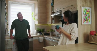 Kadın ve erkek, modern bir mutfakta rahat bir şekilde sohbet ediyorlar. Elinde kahve fincanı tutan kadınla birlikte. Parlak ve davetkar bir ortamda.