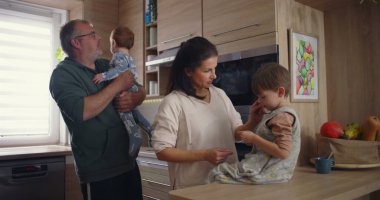 Modern mutfaktaki aile etkileşimi, baba bebeğini kucağında tutarken annesinin tezgahta oturması, sıcak aile bağlarının kurulması.