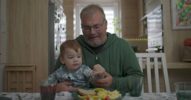 Babam yemek masasında kucağında bebeğini tutarken gülümsüyor, sıcak ve sevgi dolu bir aile anı yaratıyor.