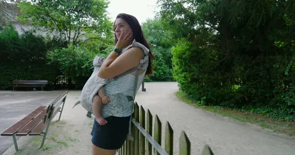 Bir taşıyıcıda bebeğini taşıyan anne, parkta dikilirken elini yüzünü dinlendirirken, bir anlık derin düşüncelere dalmış ve çevresiyle olan bağlantısını yansıtıyor.