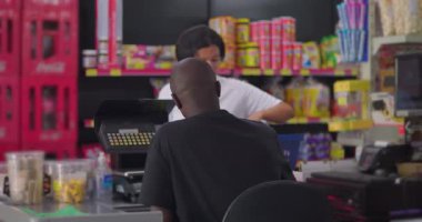 Afrikalı Amerikalı süpermarket çalışanı, arka planda renkli perakende rafları olan, günlük market sahnesi ve kasada bakkaliye alışverişi yapan Latin müşterilere yardım ediyor.