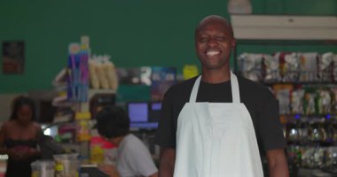 Afro-Amerikalı bir adam süpermarkette gülümsüyor, önlük giymiş neşeli bir işçi, rengarenk iç perakende ortamında güvenle duruyor.