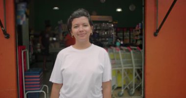 Gülümseyen Latin bir dükkan sahibi açık bir marketin önünde gururla duruyor, müşteriler için hazır girişte sıra bekliyor ve turuncu duvarlar mağazayı çerçeveliyor.