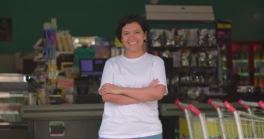 Gülümseyen Latin kadın süpermarkette kendine güvenen, kollarını kavuşturmuş, arka planda renkli ürünlerle günlük ortamda neşeli bir ifade sergiliyor.