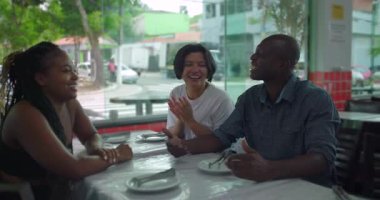 Restoran masasında gülen ve konuşan üç farklı arkadaş. İçeride neşeli ve rahat bir an. Şehir manzaralı pencerenin yanında neşeli bir sohbet.
